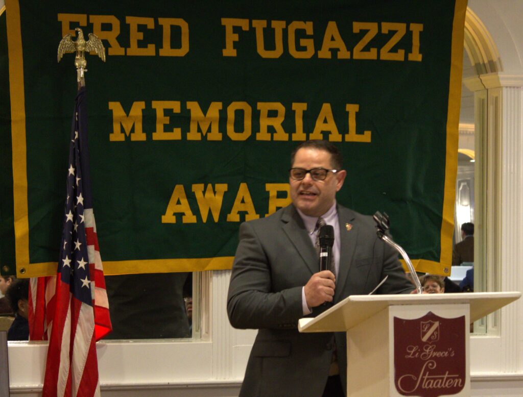 A man standing at a podium in front of a banner that says Fred Fugazi Memorial Award, showcasing his alma mater, Tottenville.