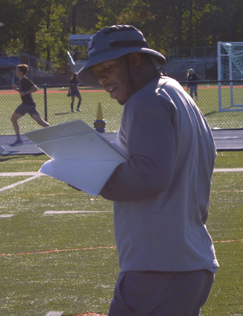 A man in athletic wear and a hat studies papers on a sports field, with road warriors running in the background, embodying the spirit of transition.