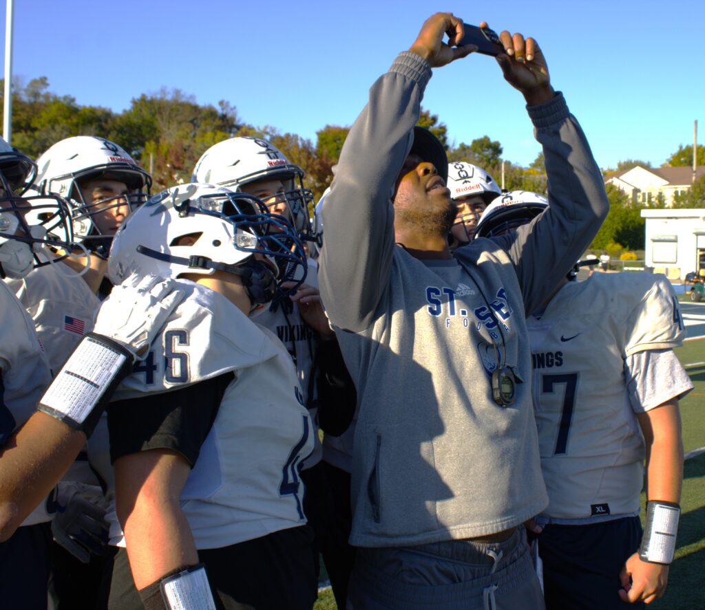 A football coach in a hoodie takes a selfie with his road warriors team on the field. Players are in white helmets and jerseys, ready for the transition to sea football. Trees and a building stand visible in the background.