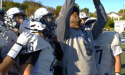 A football coach in a hoodie takes a selfie with his road warriors team on the field. Players are in white helmets and jerseys, ready for the transition to sea football. Trees and a building stand visible in the background.