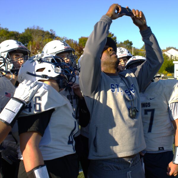 A football coach in a hoodie takes a selfie with his road warriors team on the field. Players are in white helmets and jerseys, ready for the transition to sea football. Trees and a building stand visible in the background.