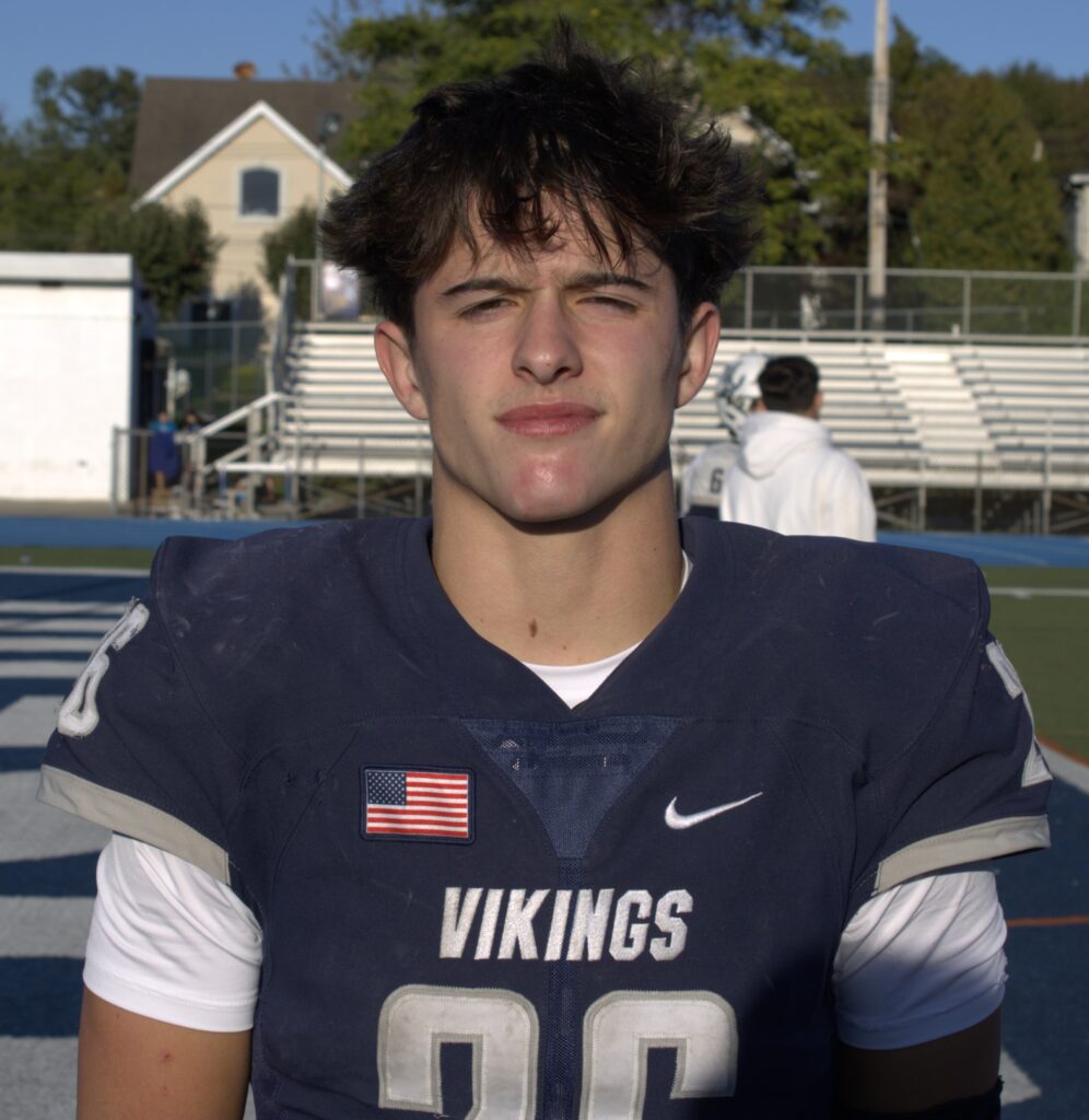 A football player donning a jersey with "Vikings" emblazoned on it stands confidently on an outdoor field. In the background, fellow road warriors prepare for the game amidst the bleachers and eager teammates.